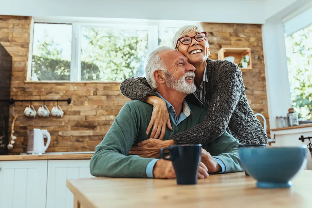Two older adults, husband and wife, sitting their kitchen together smiling and hugging